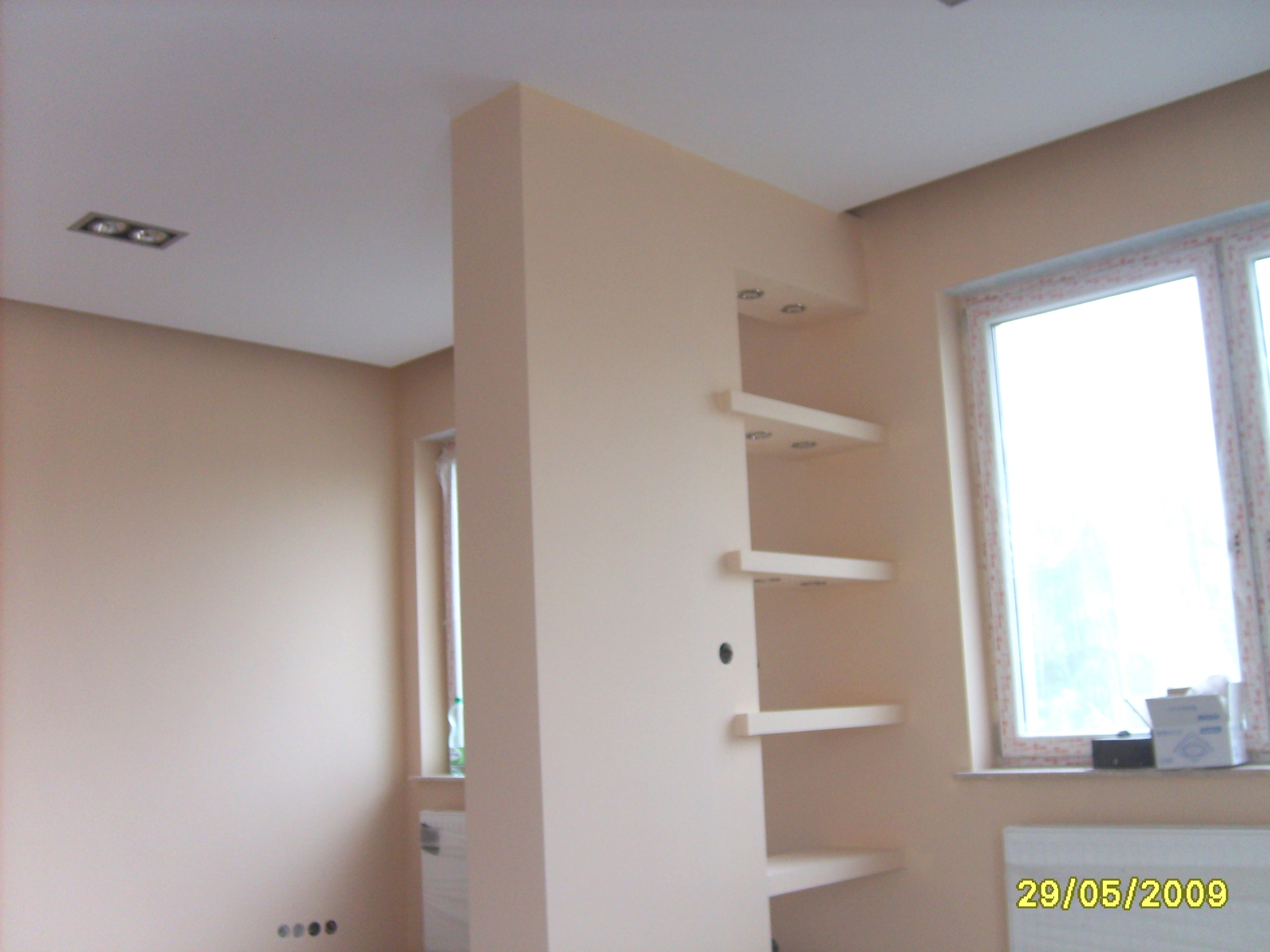 Wykończone wnętrze w jasnych odcieniach beżu, z ścianką działową z półkami i wbudowanym oświetleniem punktowym, widok na okno z parapetem.